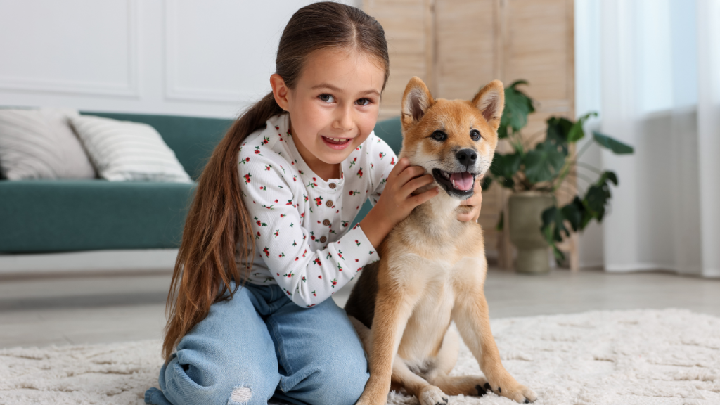 Ein lächelndes junges Mädchen sitzt auf dem Teppich und umarmt einen fröhlichen, hellbraunen Hund. Beide schauen in die Kamera.